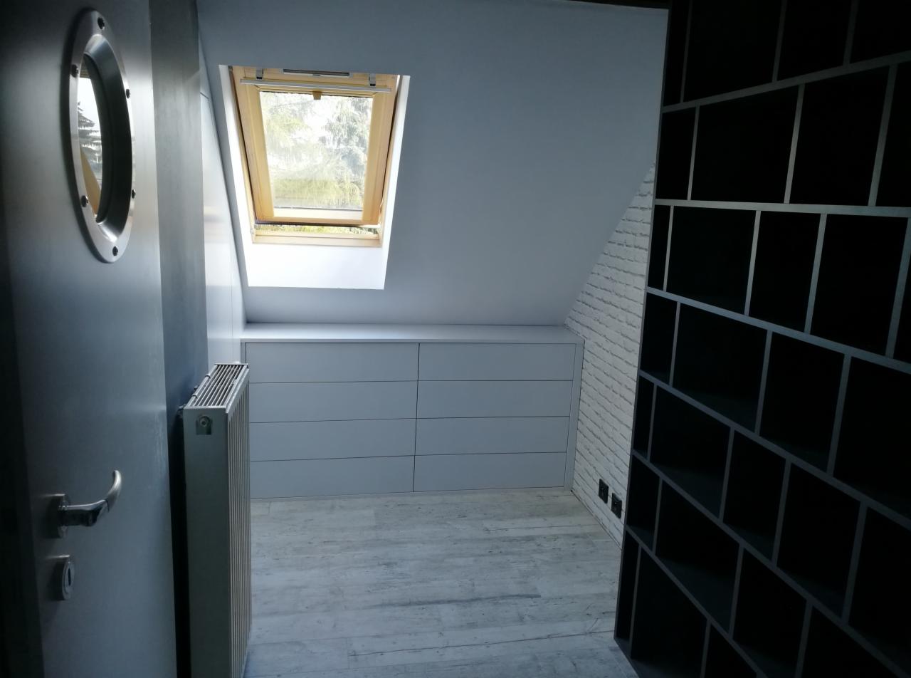 Szafa wnękowa z szufladami pod skosem dachu i czarny regał w pokoju na poddaszu w Bartkowie, widok zza drzwi z okrągłym oknem.