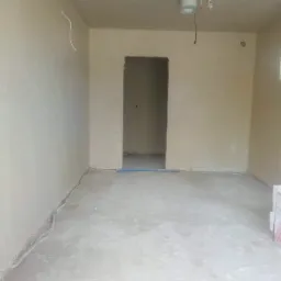 Wnętrze pomieszczenia po nałożeniu tynku maszynowego, widoczne ściany o jednolitej strukturze, sufit z zamontowaną lampą i elementami instalacji, oraz przewody elektryczne przy podłodze.