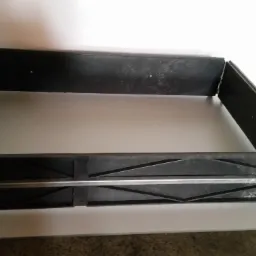 Czarna, prostokątna rama z metalowymi wzmocnieniami i narożnikami, leżąca na białej powierzchni.