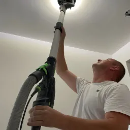 Pracownik szlifuje sufit za pomocą szlifierki teleskopowej Festool, widok z dołu, oświetlenie robocze podkreśla niedoskonałości powierzchni przygotowanej pod malowanie.