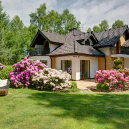 Dwupiętrowy dom z ciemnym dachem, balkonem z ozdobną balustradą i tarasem otoczony kwitnącymi rododendronami w odcieniach różu i bieli, z rattanowym zestawem wypoczynkowym na trawniku.