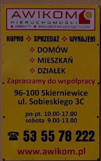 Żółty szyld agencji nieruchomości Awikom z ofertą kupna, sprzedaży i wynajmu domów, mieszkań i działek, adres Skierniewice, ul. Sobieskiego 3C, godziny otwarcia i numer telefonu.