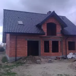 Surowy stan domu jednorodzinnego z czarnym dachem z dachówki ceramicznej, widoczne mury z czerwonej cegły, brak okien i drzwi, teren wokół domu niezagospodarowany, pochmurne niebo.