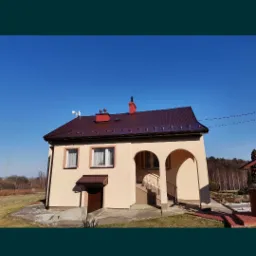 Dom jednorodzinny z beżową elewacją, brązowym dachem i czerwonymi kominami na tle błękitnego nieba.