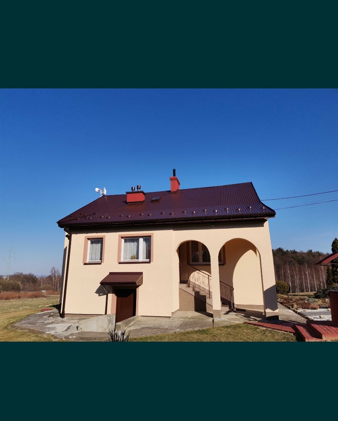 Dom jednorodzinny z beżową elewacją, brązowym dachem i czerwonymi kominami na tle błękitnego nieba.