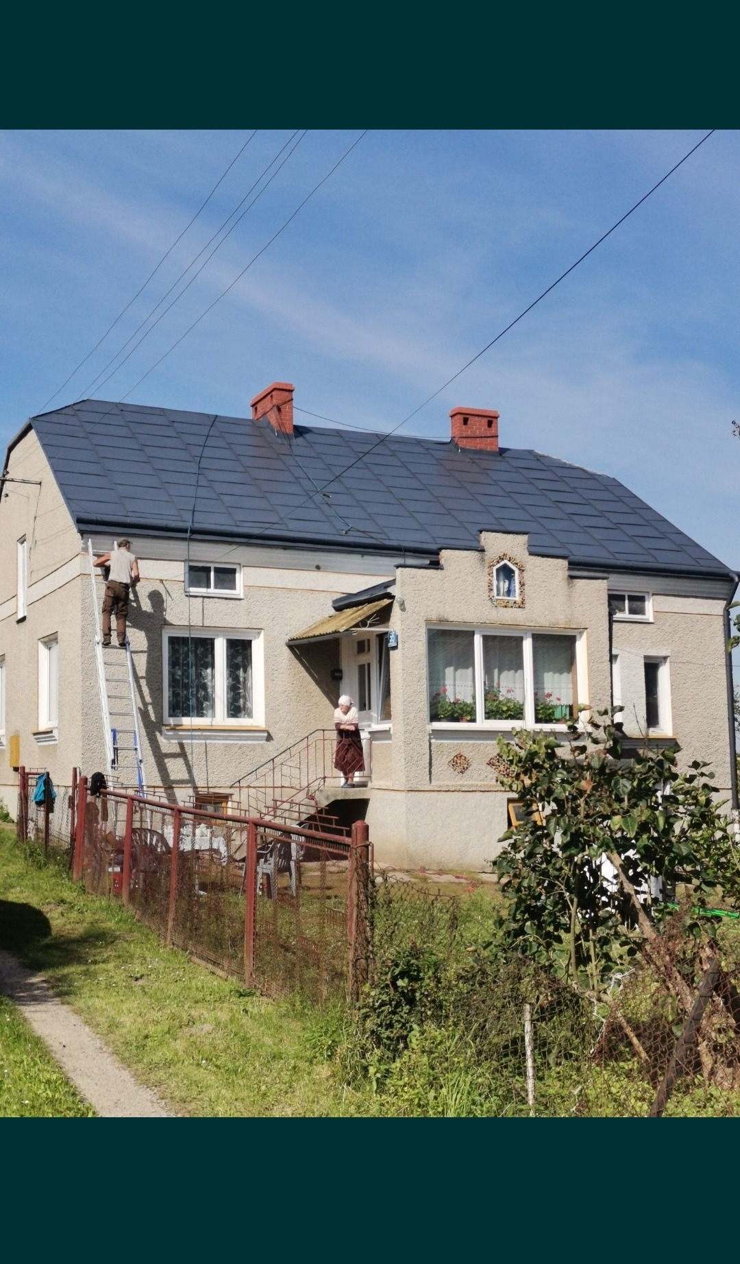 Malowanie elewacji domu jednorodzinnego z drabiny, widoczna kobieta na ganku i świeżo pomalowany dach z czerwonymi kominami.