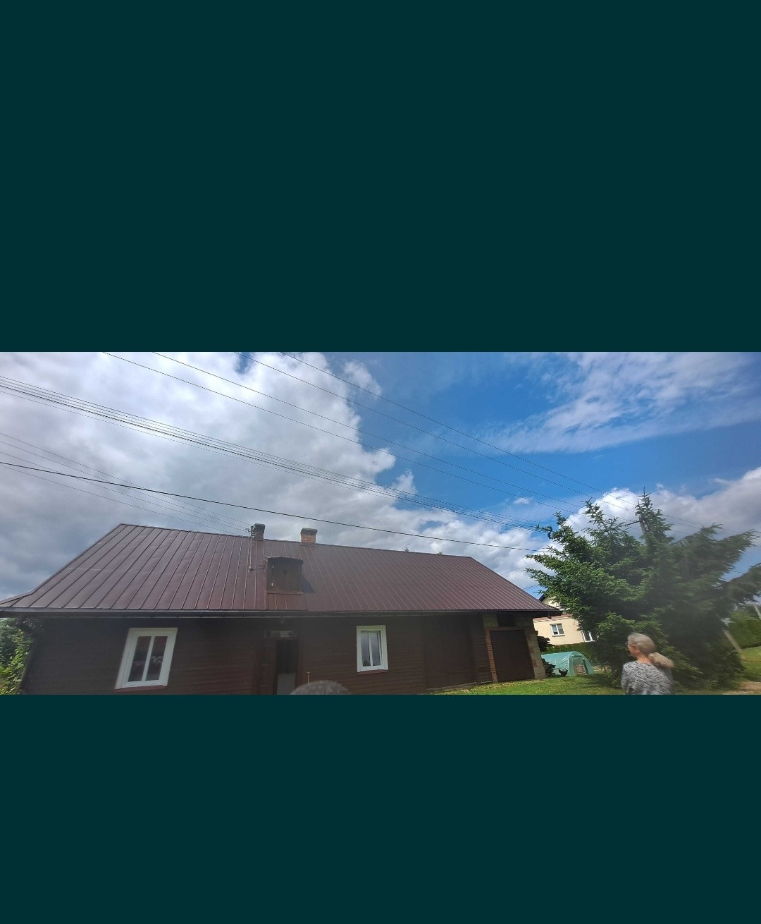 Dom z nowym, brązowym dachem z blachy trapezowej, widoczne kominy, okna i fragment ogrodu z drzewem iglastym na tle błękitnego nieba z chmurami.