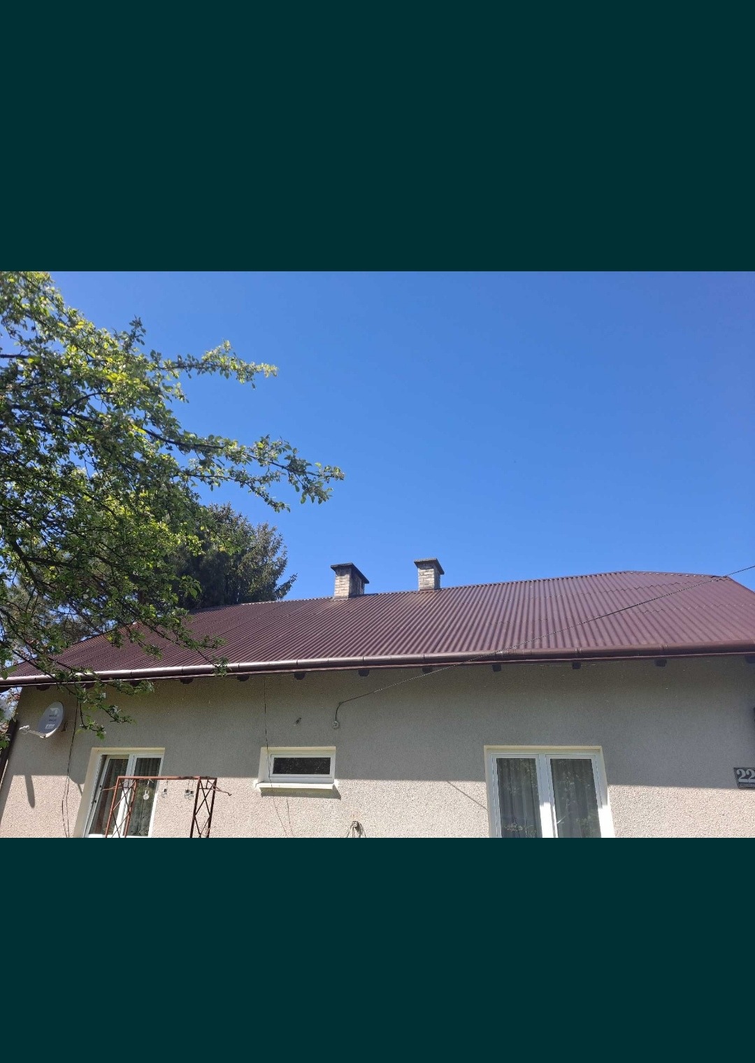 Dom jednorodzinny z nowym, brązowym dachem z blachy trapezowej i dwoma kominami, widok z dołu z fragmentem drzewa po lewej stronie.