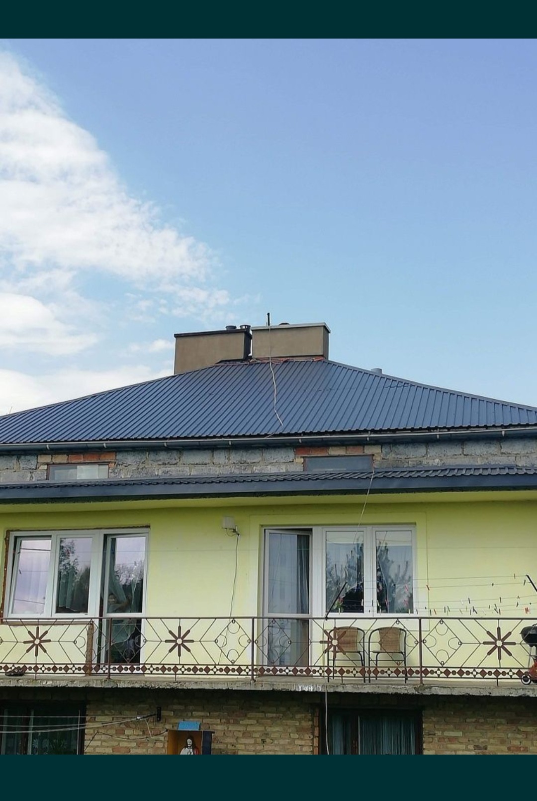 Dom z nowym, ciemnoszarym dachem z blachy trapezowej i żółtą elewacją, widok z dołu na balkon z ozdobną balustradą.