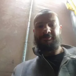 Portret brodatego mężczyzny w roboczym ubraniu na tle ściany z widoczną instalacją elektryczną.