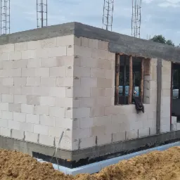 Surowy stan domu z białych bloczków, widoczne stalowe pręty zbrojeniowe wystające z górnej części ścian, fundament ocieplony styropianem, wykopana ziemia wokół budynku.