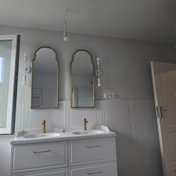 GrzesDeko Profesjonalne Wykończenia Wnętrz - Jasna łazienka z dwoma lustrami w złotych ramach nad umywalkami. Widoczne białe szafki, dekoracyjne oświetlenie i otwarte okno z widokiem na miasto.