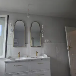 Jasna łazienka z dwoma lustrami w złotych ramach nad umywalkami. Widoczne białe szafki, dekoracyjne oświetlenie i otwarte okno z widokiem na miasto.