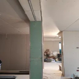 Wnętrze w trakcie montażu płyt kartonowo-gipsowych: widoczne metalowe profile sufitowe, pionowy słup obłożony zielonymi płytami, przewody elektryczne wystające ze ściany, narzędzia na podłodze.