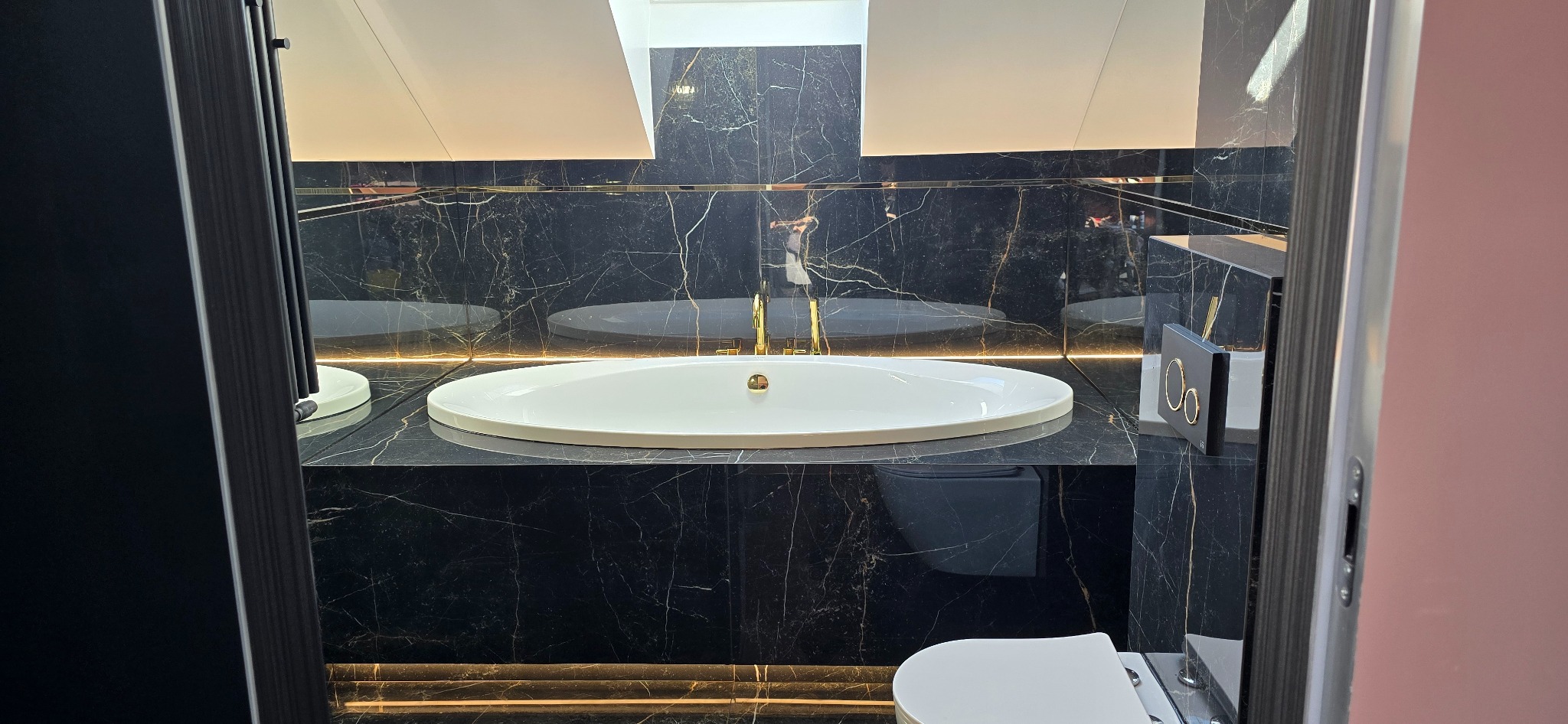 Elegancka łazienka z czarnymi marmurowymi płytkami, podświetlanym blatem, owalną umywalką i złotą armaturą, widziana zza uchylonych drzwi.