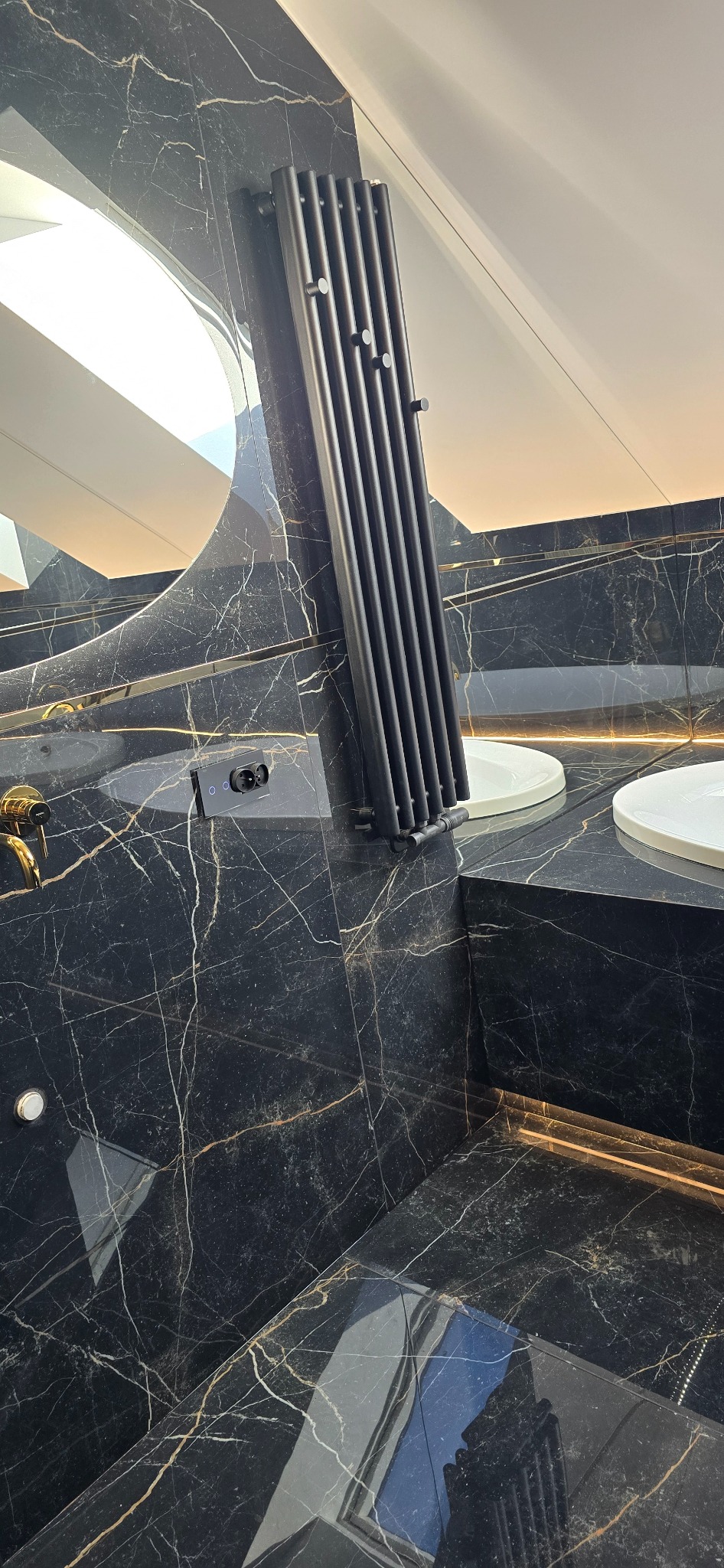 Nowoczesna łazienka z czarnymi marmurowymi płytkami, grzejnikiem drabinkowym w kolorze czarnym, podświetlanym lustrem i umywalkami nablatowymi.