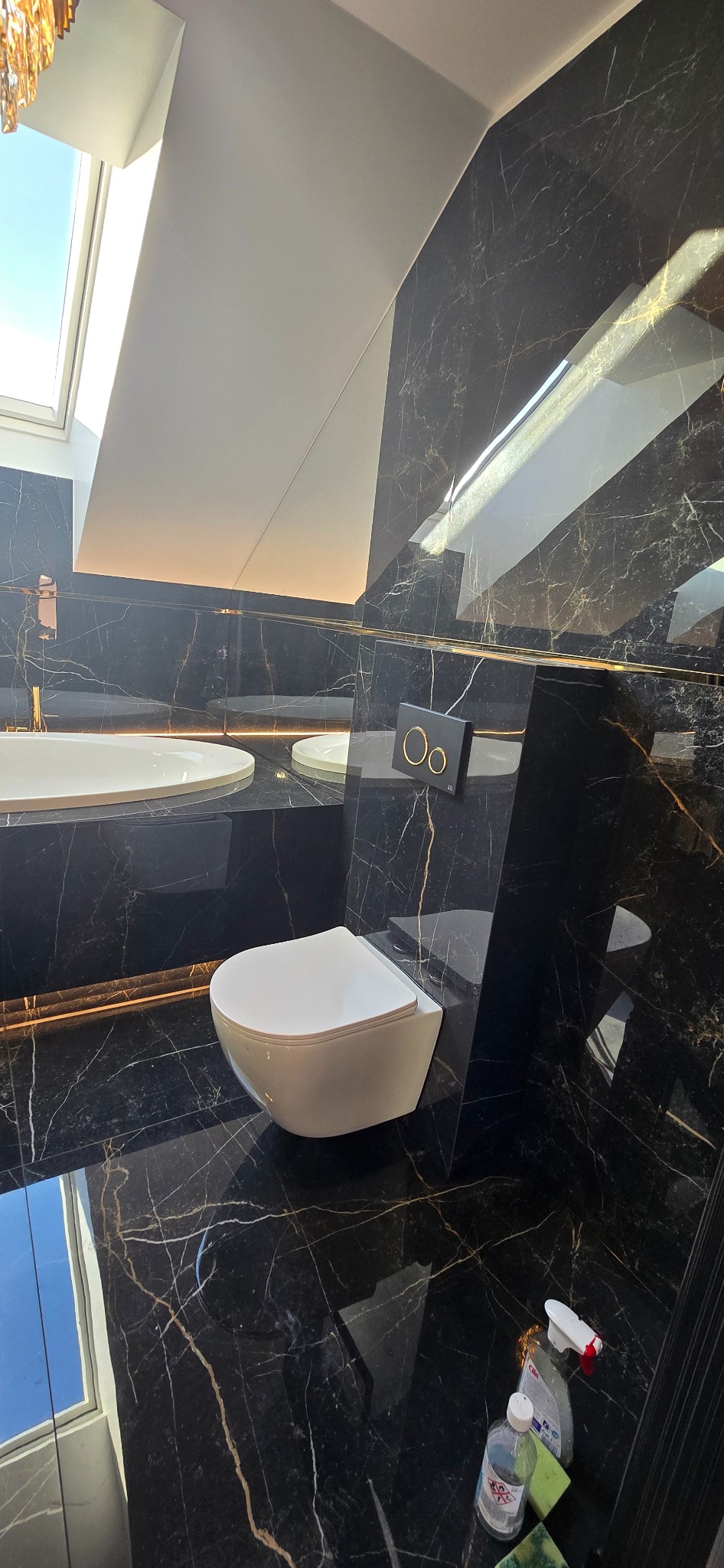 Nowoczesna łazienka z czarnymi marmurowymi płytkami, podwieszaną toaletą i oknem dachowym; widoczne środki czyszczące na podłodze.