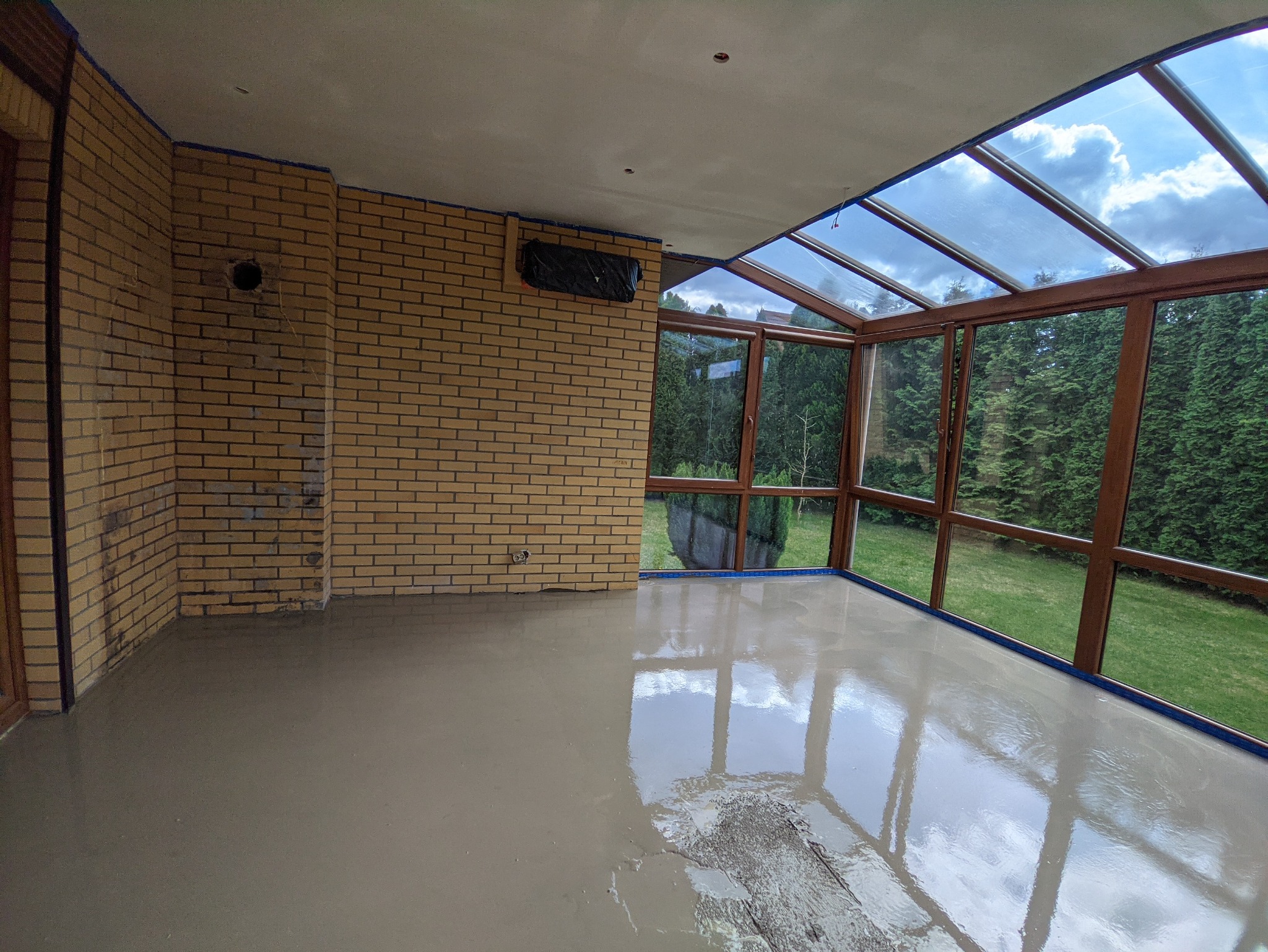 Świeżo wylana, samopoziomująca wylewka podłogowa w oranżerii z ceglaną ścianą, zabezpieczona taśmą malarską przy oknach i ścianach.