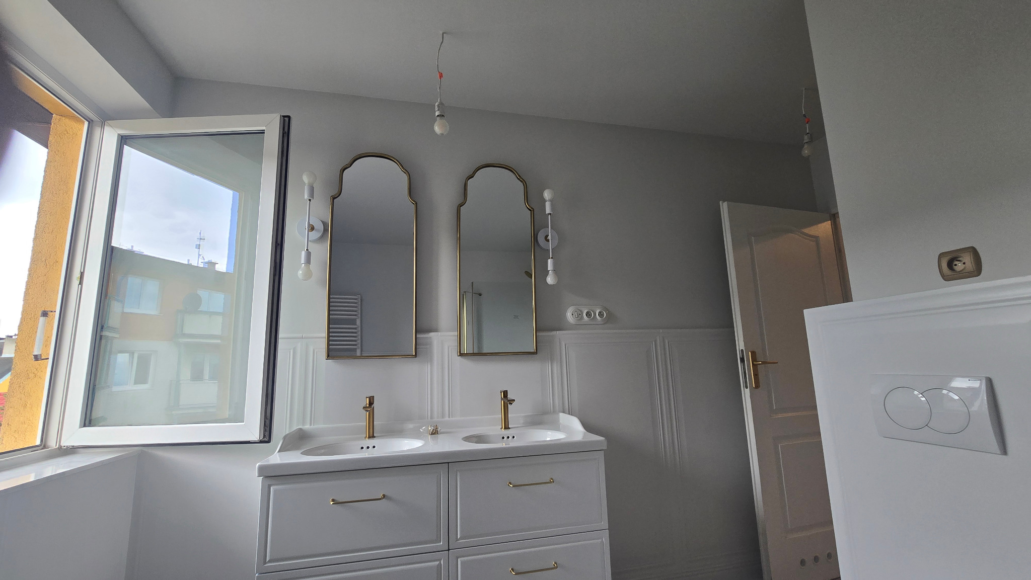 Jasna łazienka z dwoma lustrami w złotych ramach nad umywalkami. Widoczne białe szafki, dekoracyjne oświetlenie i otwarte okno z widokiem na miasto.