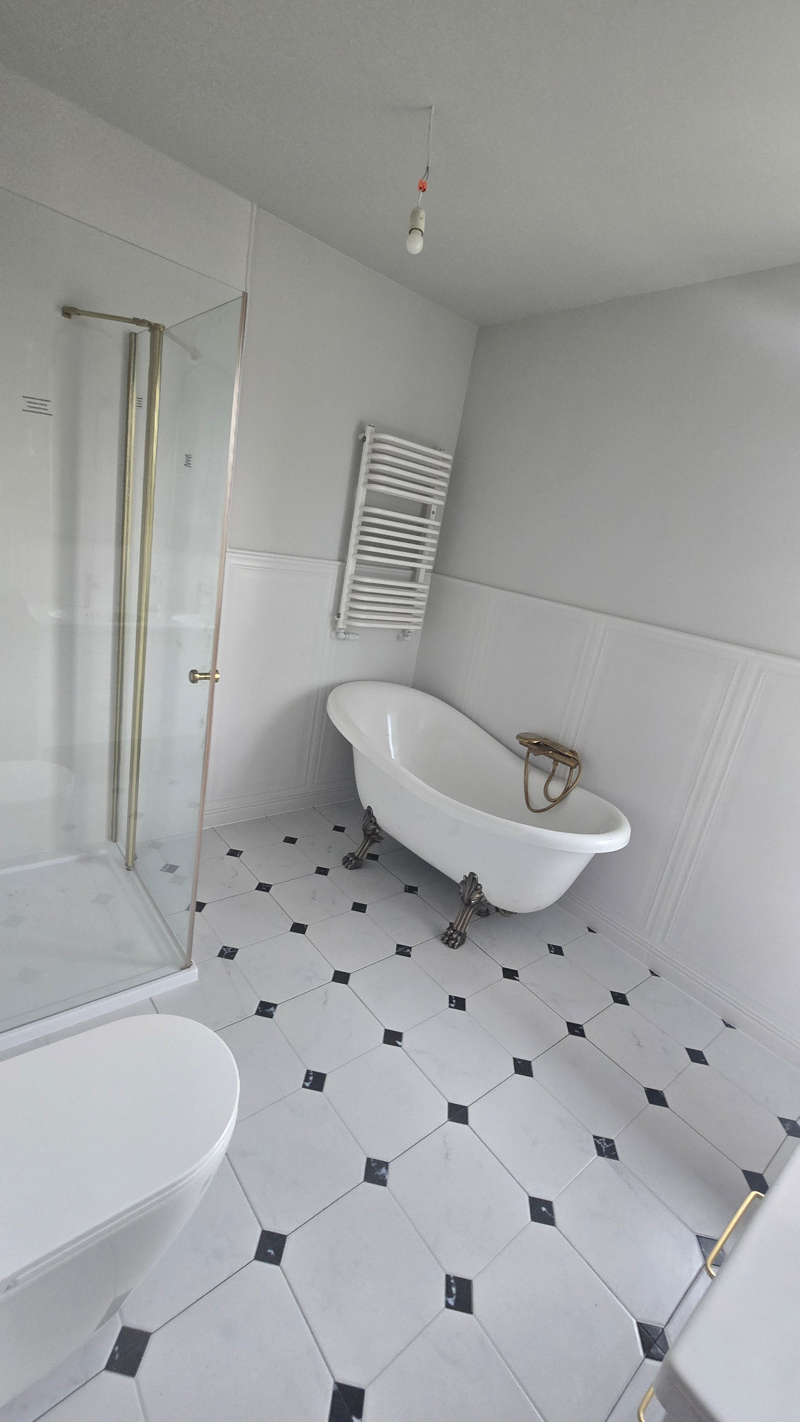 Jasne wnętrze łazienki z wolnostojącą wanną na zdobionych nóżkach, kabiną prysznicową i grzejnikiem drabinkowym. Na podłodze białe płytki z czarnymi akcentami.