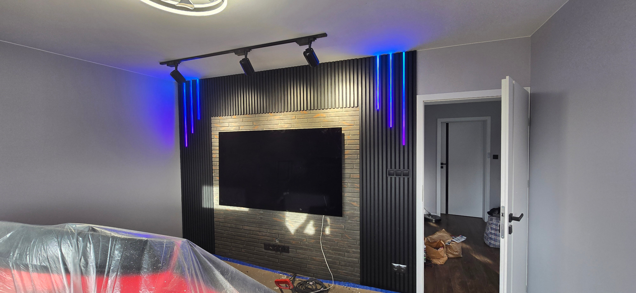 Wykończona ściana TV z panelami dekoracyjnymi, podświetleniem LED i imitacją cegły. Widoczne oświetlenie szynowe i częściowo zakryte meble folią ochronną.