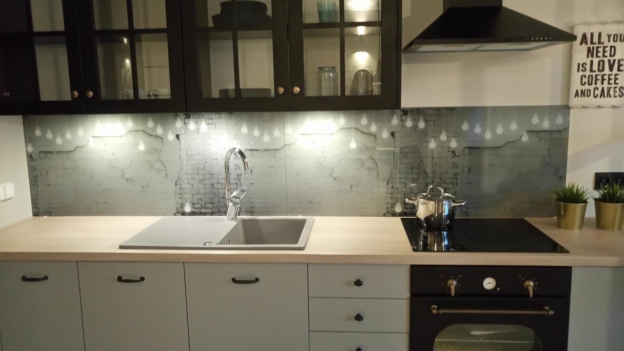 Nowoczesna kuchnia z szarymi szafkami, drewnianym blatem i szklanym panelem z motywem cegieł i żarówek, z czarnym okapem i piekarnikiem w stylu retro.