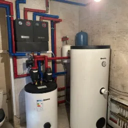 Zestaw dwóch białych zbiorników akumulacyjnych z czarnymi pokrywami, połączonych rurami w kolorach czerwonym i niebieskim, z widocznymi elementami instalacji i wiszącą żarówką w tle.