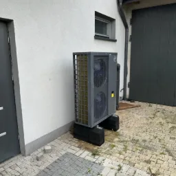 Szara pompa ciepła zamontowana na czarnych podstawach przy ścianie budynku w Rząśni, widoczne dwa wentylatory wewnątrz obudowy i kostka brukowa z chwastami.