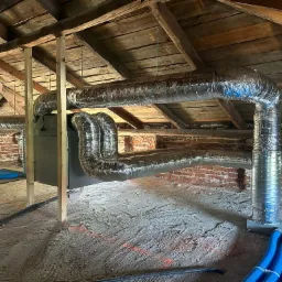 Instalacja rekuperacji na poddaszu: srebrne, elastyczne rury wentylacyjne pod drewnianym stropem, widoczny centralny moduł rekuperatora i niebieskie przewody na podłodze z surowego betonu.