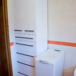 Biała szafa łazienkowa z szufladami i szafkami, z długimi uchwytami, stoi obok pralki w jasnej łazience z pomarańczowym akcentem na płytkach.