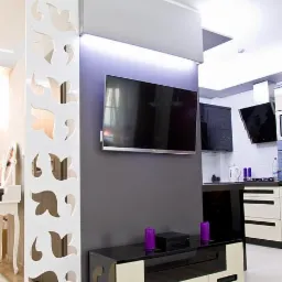 Nowoczesny salon z ażurową ścianką działową, szafką RTV i telewizorem. W tle widoczna kuchnia z czarnymi frontami i białymi elementami. Minimalistyczny wystrój.