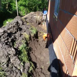 Fundament budynku w trakcie izolacji, widoczny wykop, folia izolacyjna i mur z pustaków ceramicznych. W tle pracownik z łopatą.