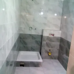 Nowoczesna łazienka z szaro-białymi płytkami imitującymi marmur, kabina prysznicowa w rogu, widoczne przygotowane przyłącza wodno-kanalizacyjne.