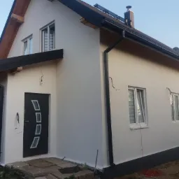 Dom jednorodzinny z nową, jasną elewacją, czarnymi elementami wykończeniowymi dachu i okien, widoczne ślady po przewodach elektrycznych przy oknach.