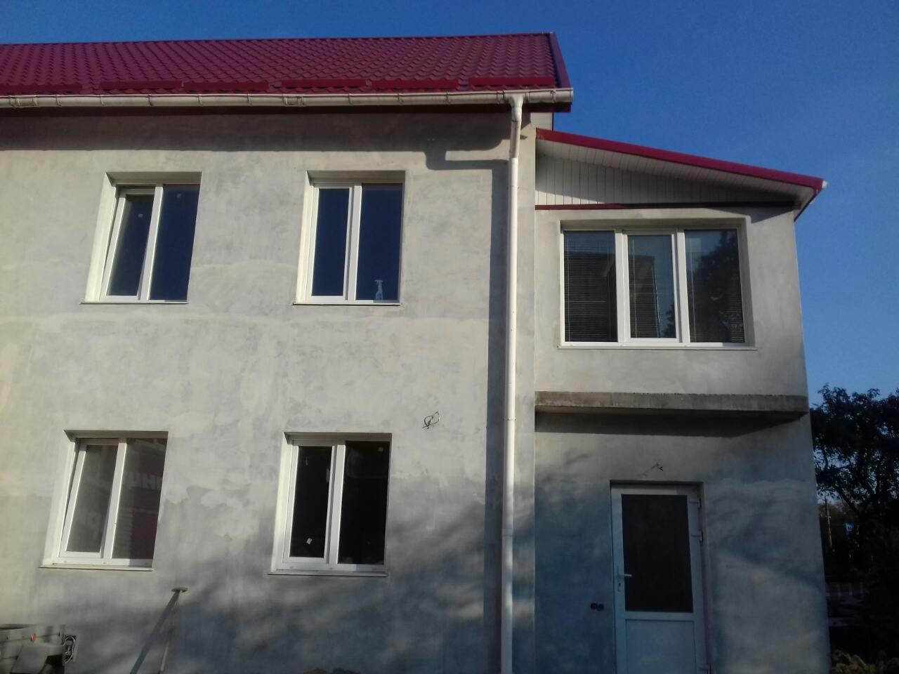 Dwukondygnacyjny dom w trakcie prac elewacyjnych, surowy tynk szary, czerwony dach, biała stolarka okienna i drzwiowa, rynna spustowa z boku budynku.