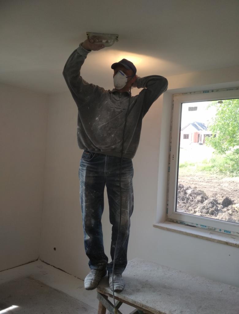 Pracownik w masce, stojący na podwyższeniu, wygładza sufit szpachelką w nowym domu z widokiem na ogród przez okno.