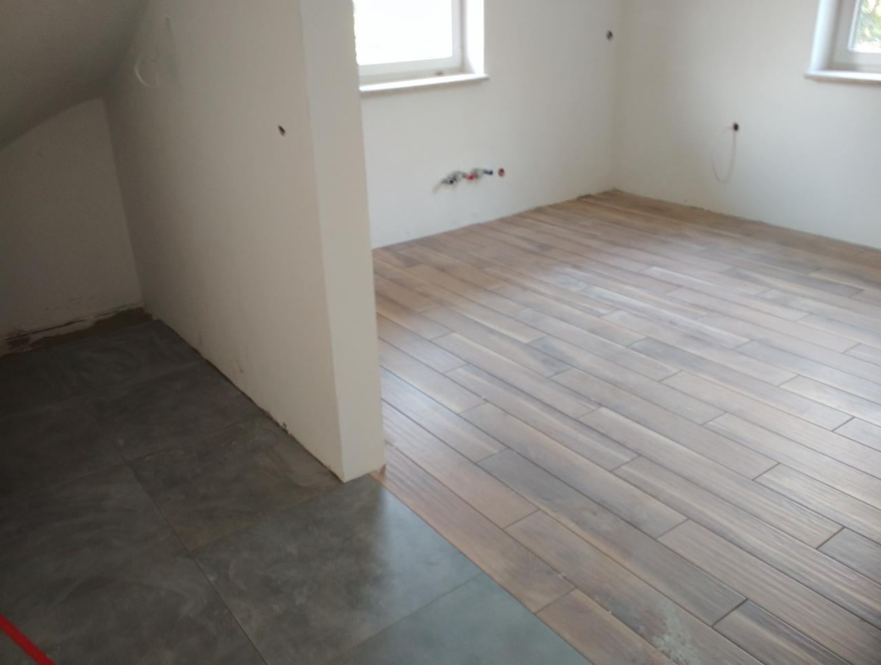 Dwa rodzaje płytek podłogowych w łazience: imitacja drewna i szare płytki, widoczne instalacje na ścianie, surowy stan wykończenia.