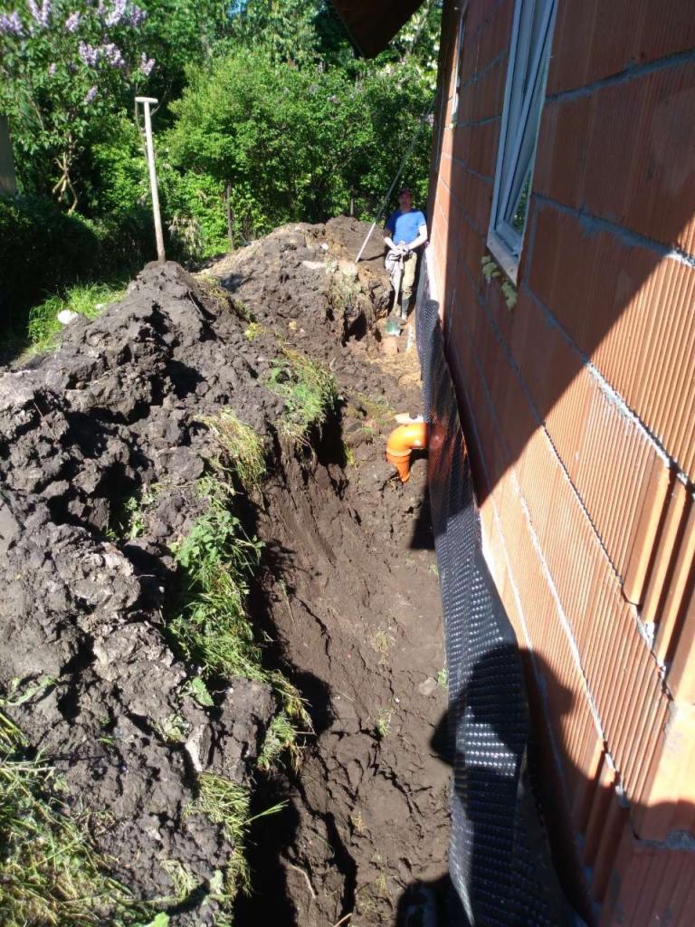 Fundament budynku w trakcie izolacji, widoczny wykop, folia izolacyjna i mur z pustaków ceramicznych. W tle pracownik z łopatą.
