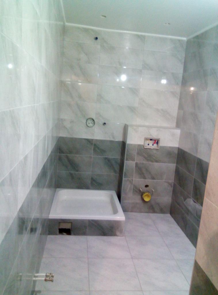 Nowoczesna łazienka z szaro-białymi płytkami imitującymi marmur, kabina prysznicowa w rogu, widoczne przygotowane przyłącza wodno-kanalizacyjne.