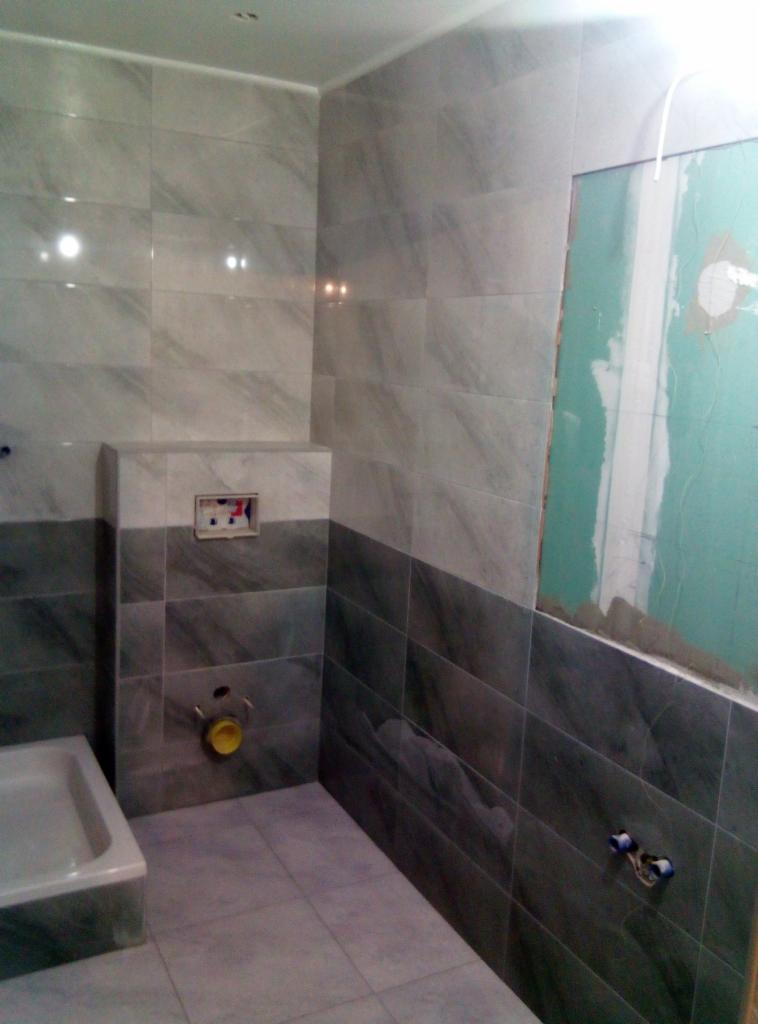 Układanie płytek w łazience, widoczne szare i białe płytki imitujące marmur, przygotowane instalacje wodne i fragment ściany z płyt gipsowo-kartonowych przed wykończeniem.