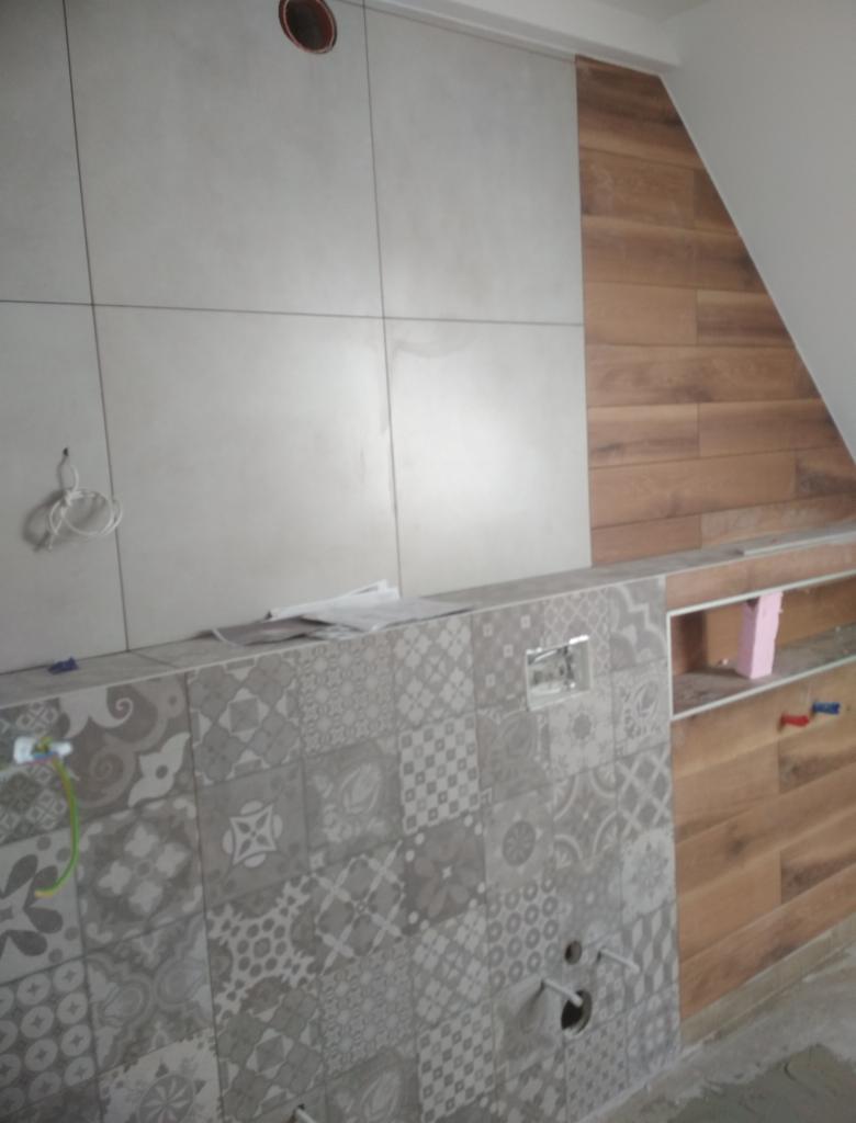 Montaż płytek w łazience: połączenie gładkich, szarych płytek wielkoformatowych z dekoracyjnymi, wzorzystymi płytkami w odcieniach szarości oraz drewnopodobnymi panelami na skośnej ścianie.