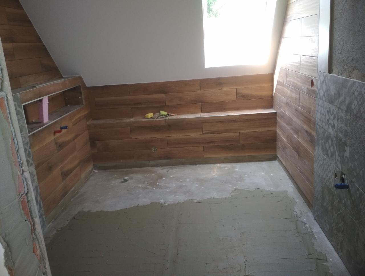 Łazienka na poddaszu w trakcie wykończenia: płytki imitujące drewno na ścianach, przygotowane podłoże pod posadzkę, widoczny fragment wnęki z instalacją.