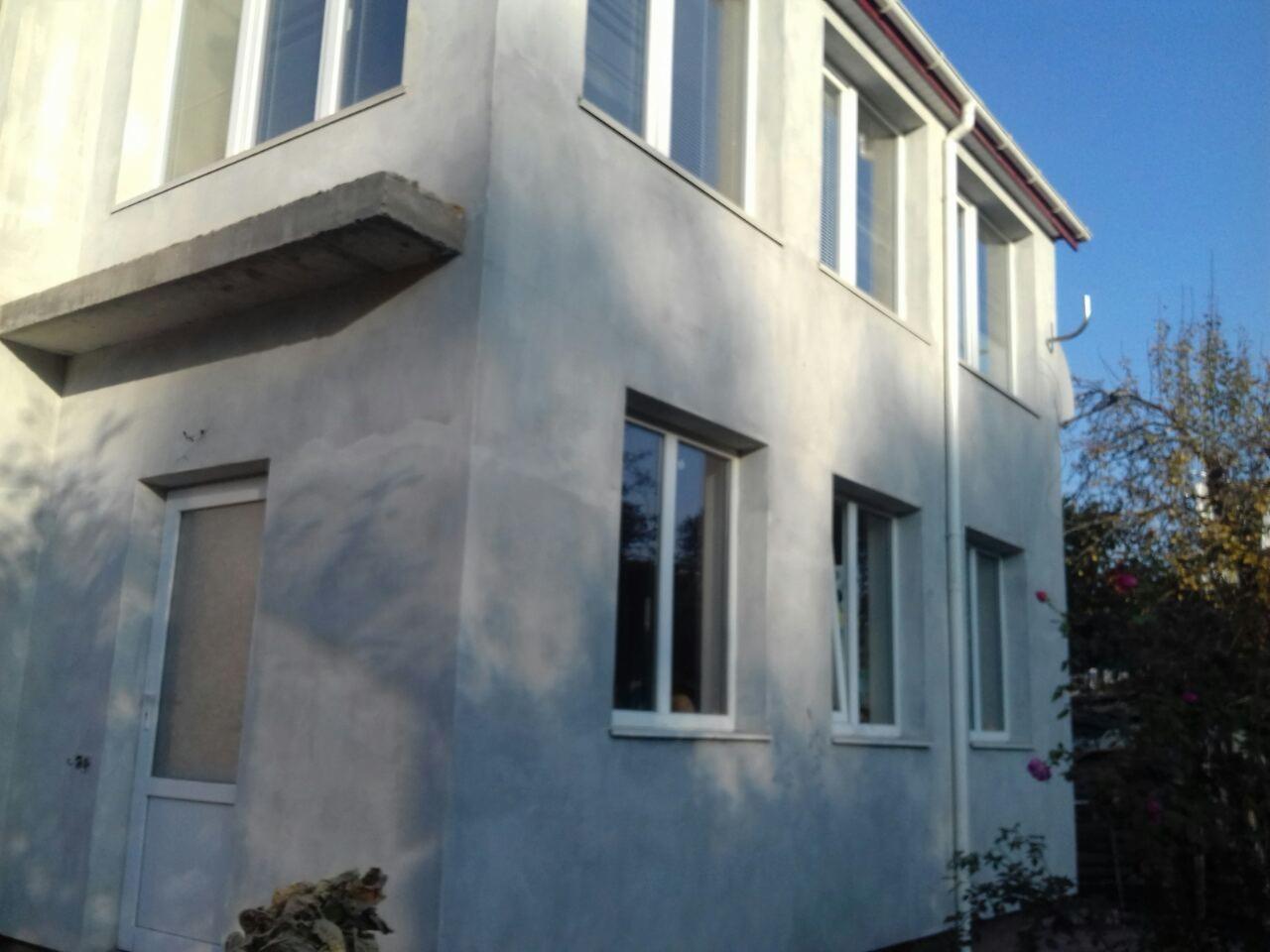 Dwupiętrowy budynek z szarą elewacją w trakcie prac wykończeniowych, widoczne okna, drzwi wejściowe i betonowy daszek nad nimi, na tle błękitnego nieba i drzew