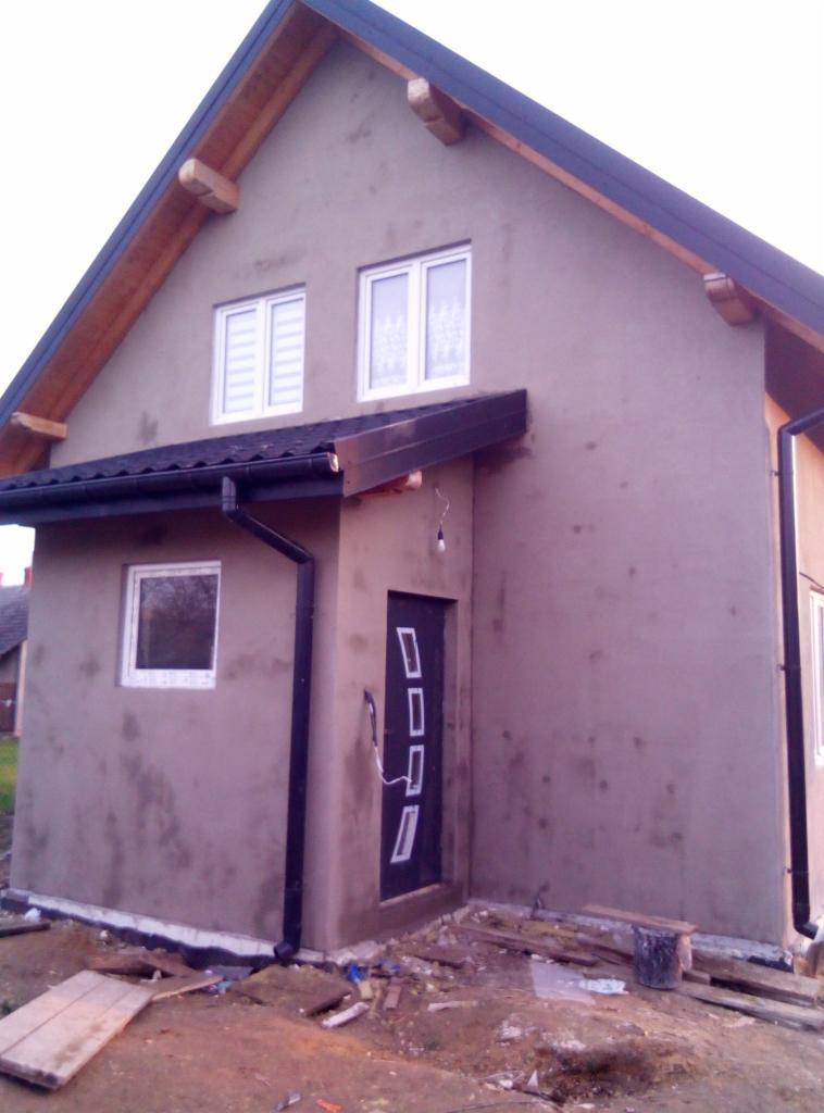 Dom jednorodzinny w trakcie nakładania szarego tynku zewnętrznego, widoczne nowe okna z białymi ramami, czarne rynny i drzwi wejściowe z pionowymi, białymi prostokątami, teren wokół domu częściowo...