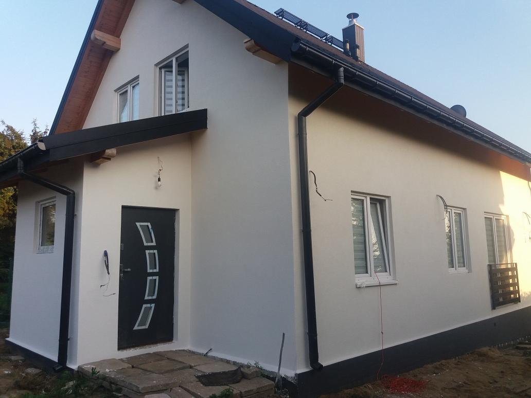 Dom jednorodzinny z nową, jasną elewacją, czarnymi elementami wykończeniowymi dachu i okien, widoczne ślady po przewodach elektrycznych przy oknach.