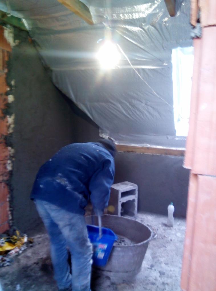 Pracownik w trakcie przygotowywania zaprawy tynkarskiej w wiadrze na budowie, ściana częściowo otynkowana, widoczna folia ochronna na poddaszu, pustak betonowy i butelka plastikowa.