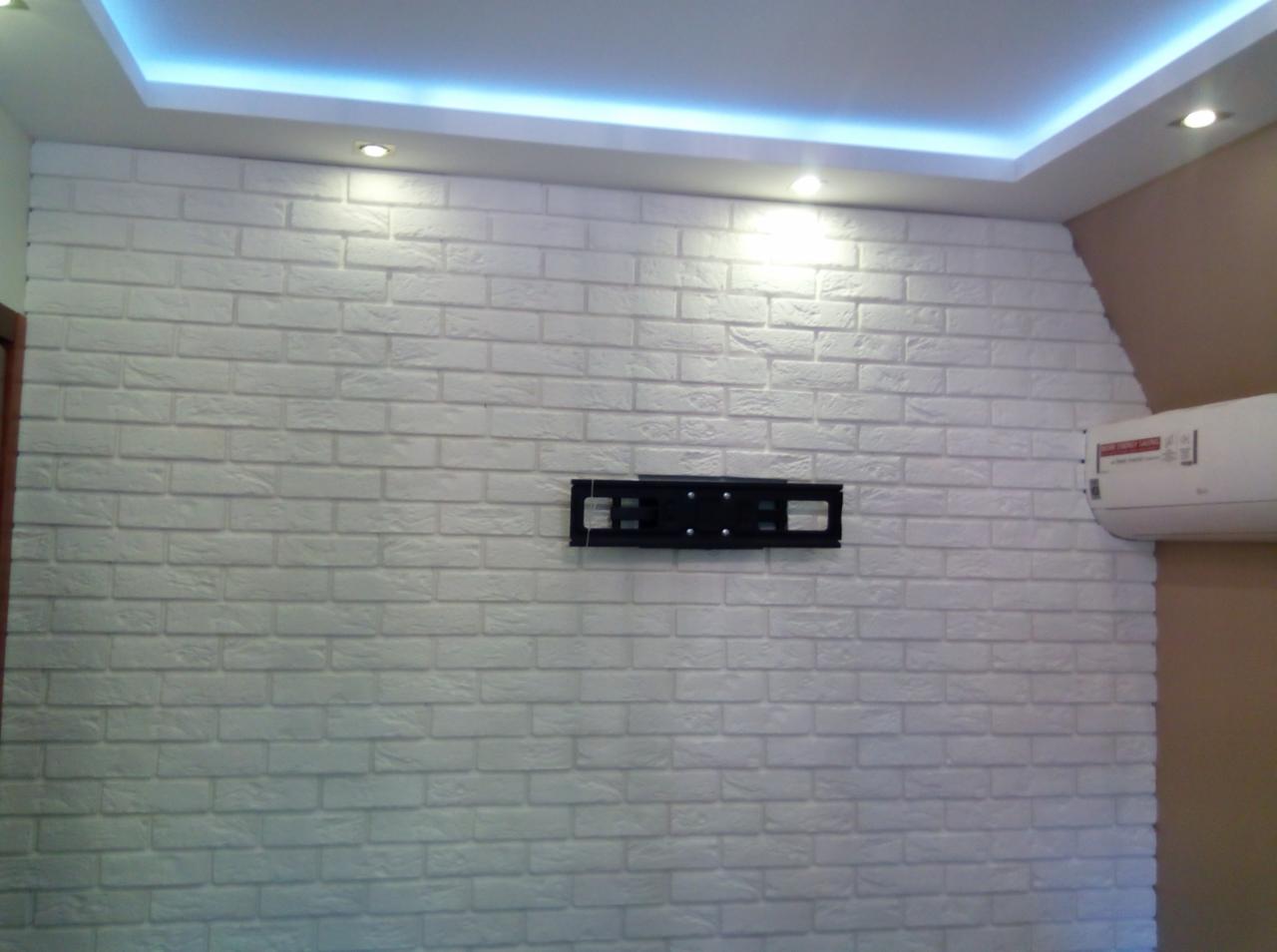 Ściana z białej cegły dekoracyjnej w salonie z zamontowanym uchwytem na telewizor i klimatyzatorem, oświetlenie sufitowe LED.