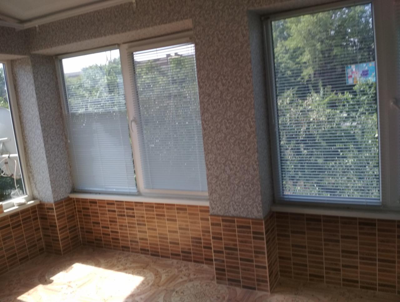 Wnętrze przeszklonego balkonu z brązowymi płytkami na ścianach i podłodze, okna z żaluzjami, tapeta w drobny wzór.