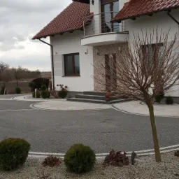 Podjazd do domu wyłożony szarą kostką brukową z białymi obrzeżami, widoczne krzewy i drzewka, dom z balkonem w tle.