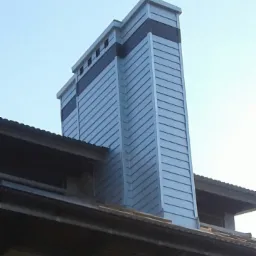 Obróbka blacharska komina z szarej blachy trapezowej, z ciemniejszymi poziomymi pasami na szczycie, widoczna z dołu, na tle jasnego nieba.